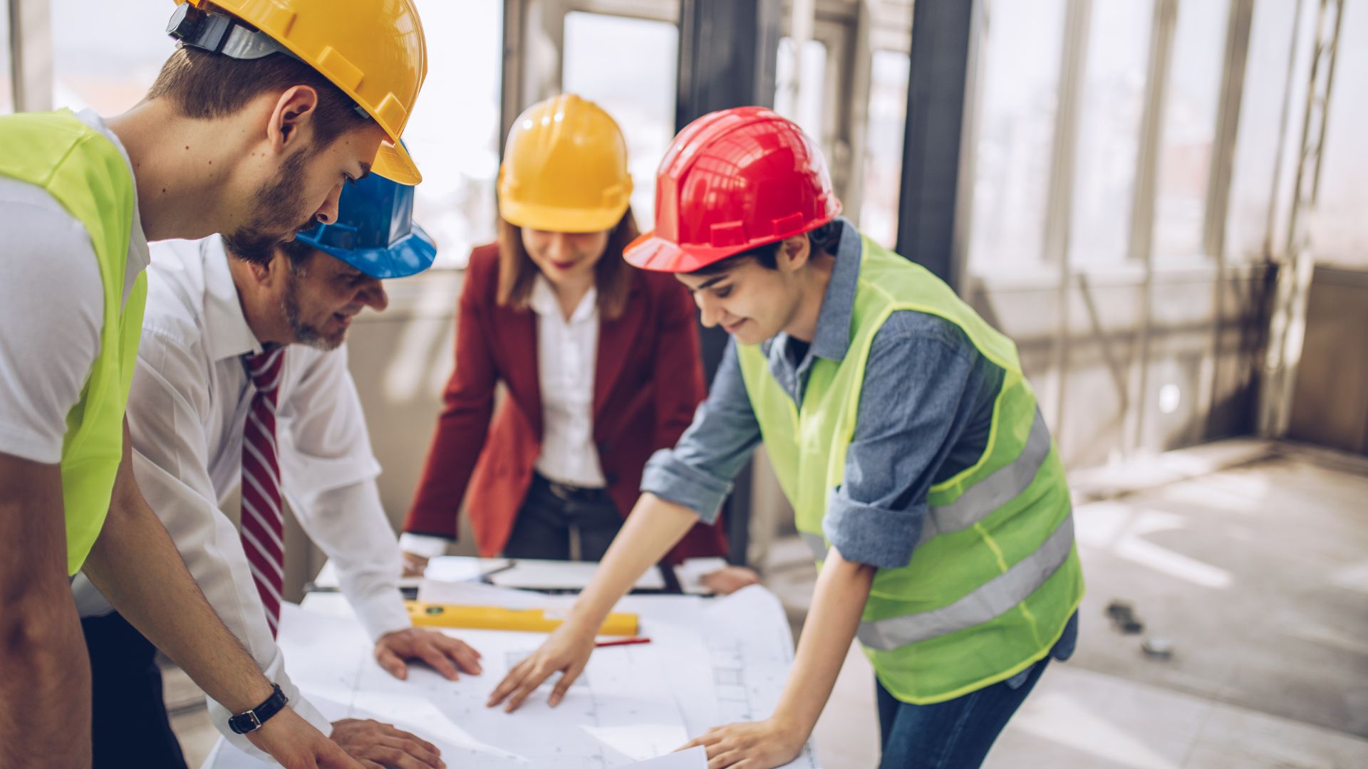 Grupo de arquitectos e ingenieros con cascos de seguridad revisando planos de construcción en una obra, trabajando en equipo dentro de un edificio en desarrollo.
