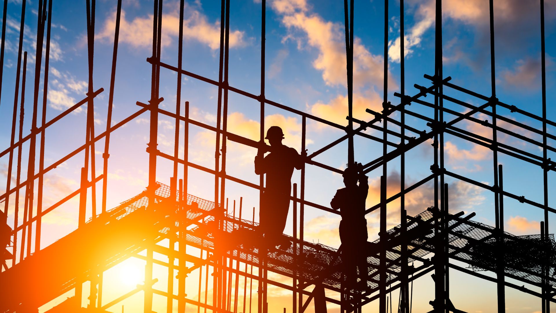: Obreros trabajando sobre andamios en una obra de construcción al atardecer, con estructura metálica en silueta y cielo azul con nubes de fondo, simbolizando proyectos en ejecución y seguridad en la obra.