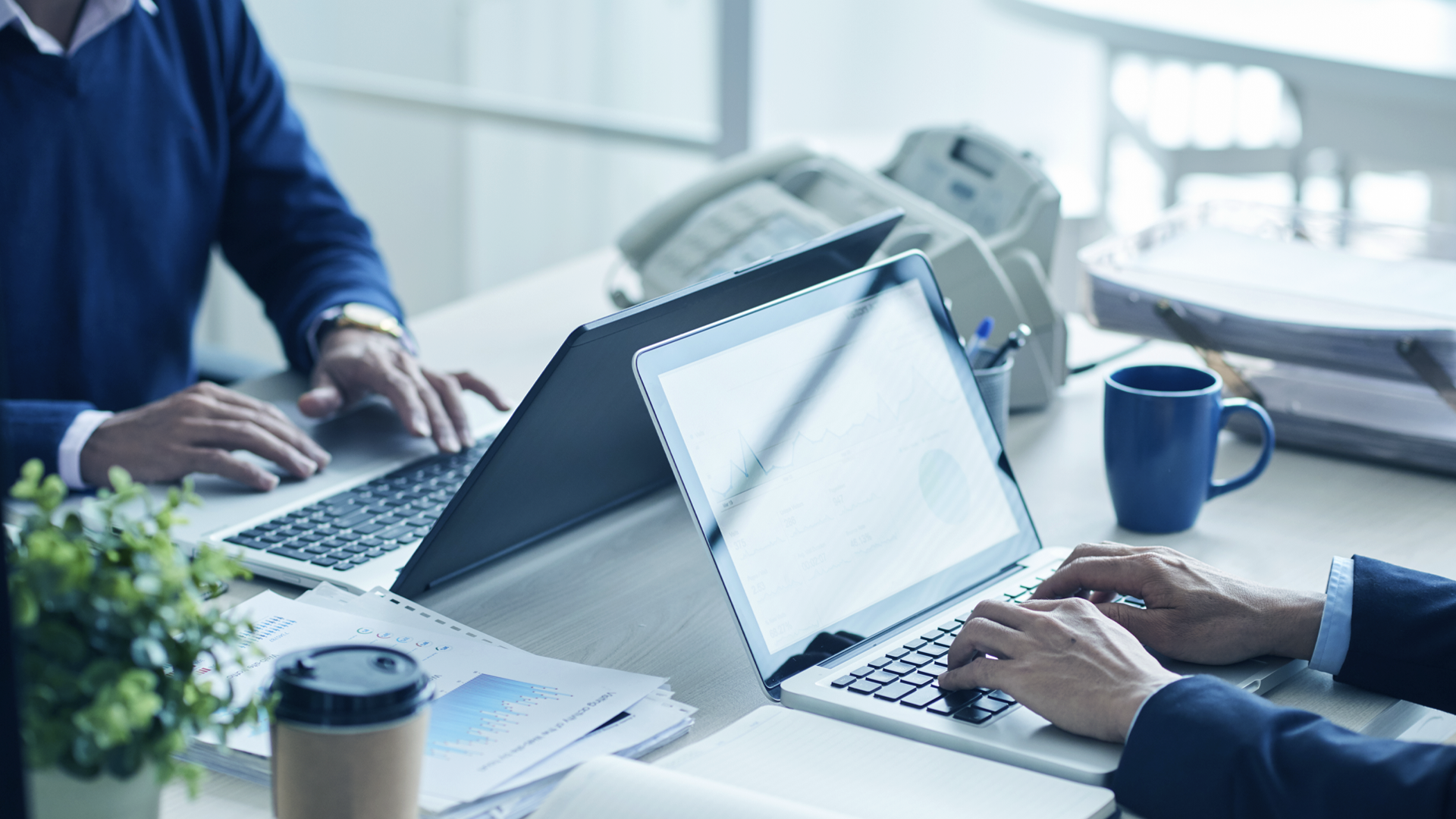 Dos personas trabajando en una oficina moderna, sentadas frente a sus computadoras portátiles. En la mesa hay papeles con gráficos, una taza de café para llevar, una planta y una taza azul. Al fondo, se observan teléfonos de oficina y una bandeja con documentos. La imagen transmite un entorno profesional y colaborativo.