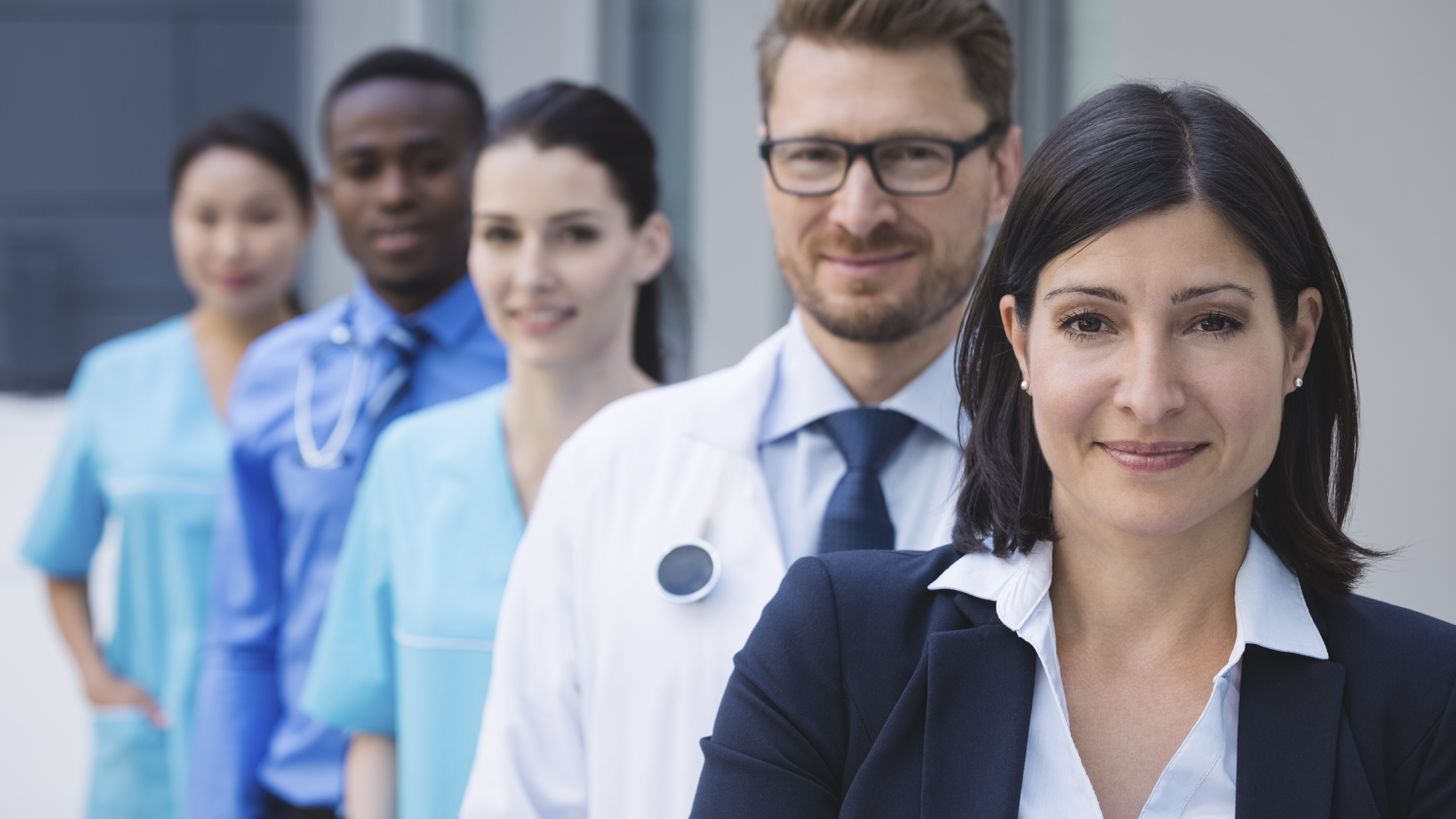 Grupo de profesionales de la salud y una mujer ejecutiva al frente, todos sonriendo y posando en fila con un fondo clínico moderno.