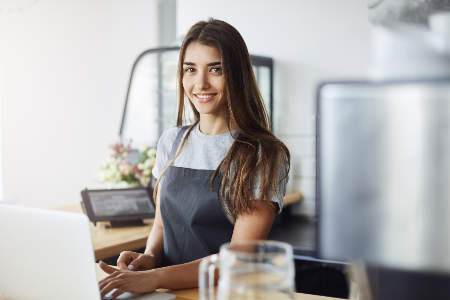 Una joven con cabello largo y castaño lleva una camiseta gris y un delantal oscuro mientras trabaja en un mostrador. Está de pie frente a una computadora portátil en lo que parece ser una cafetería o panadería, con una máquina de café y flores en el fondo.